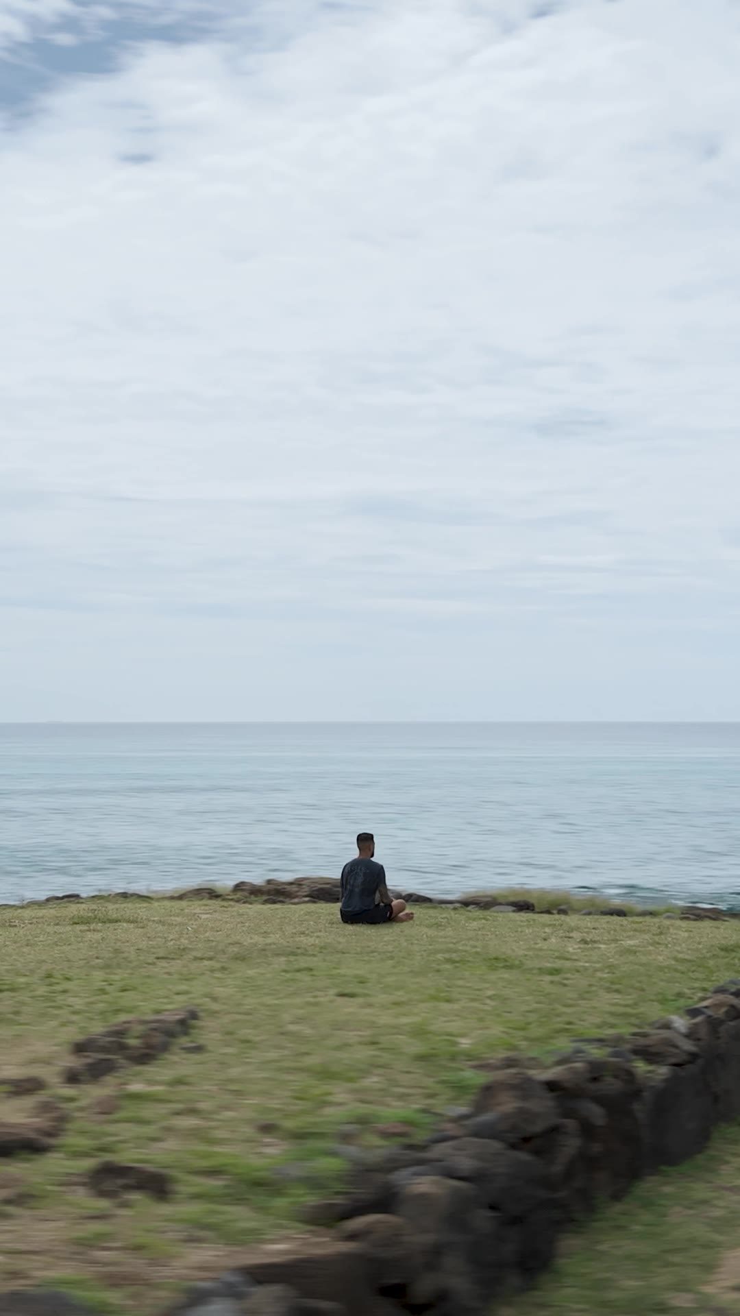 Yancy Medeiros seated on a grassy point facing the ocean on Oʻahu
